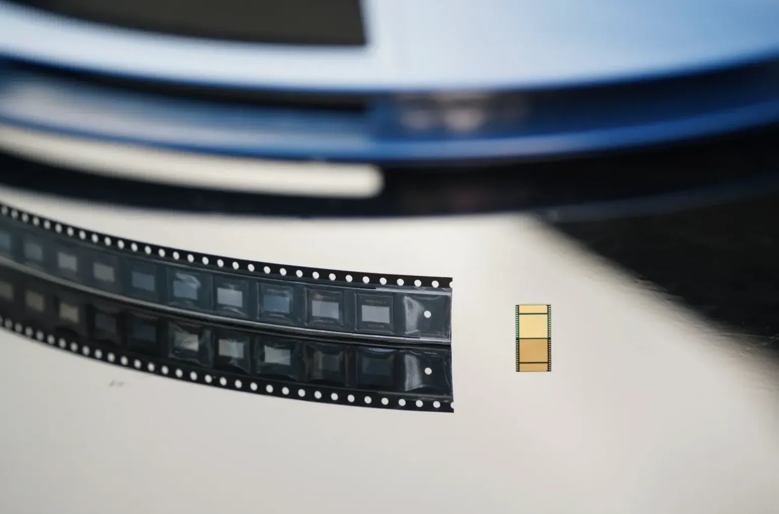 Nærbilde av en stripe bæretape med flere BM1370 ASIC-brikker, med én enkelt rektangulær brikke ved siden av som viser undersiden med gullbelagte kontaktpunkter.